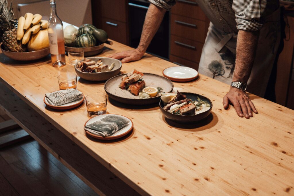 Crop unrecognizable male leaning on wooden desk with delicious appetizers and fresh fruits against alcoholic drink in glasses at home